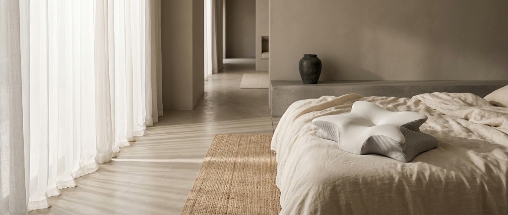 Chambre claire au sol de béton ciré : rideau de voile, tapis de jute, et l’oreiller posé sur les draps de lin d’un lit bas au second plan.