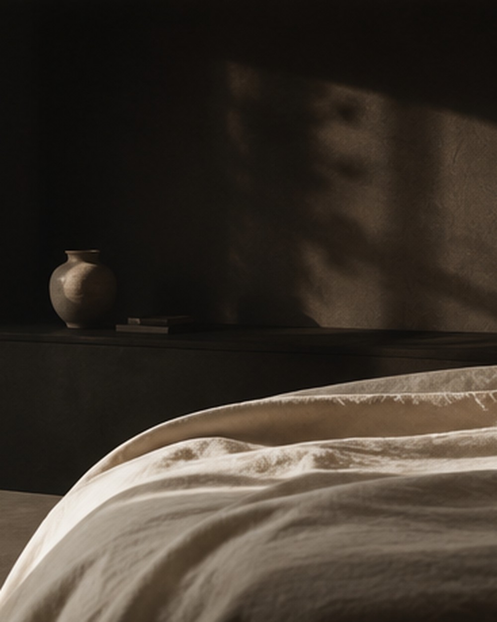 Clair-obscur dans une chambre : mur sombre où se dessine l’ombre d’un feuillage, vase en terre, couette de lin beige au premier plan.