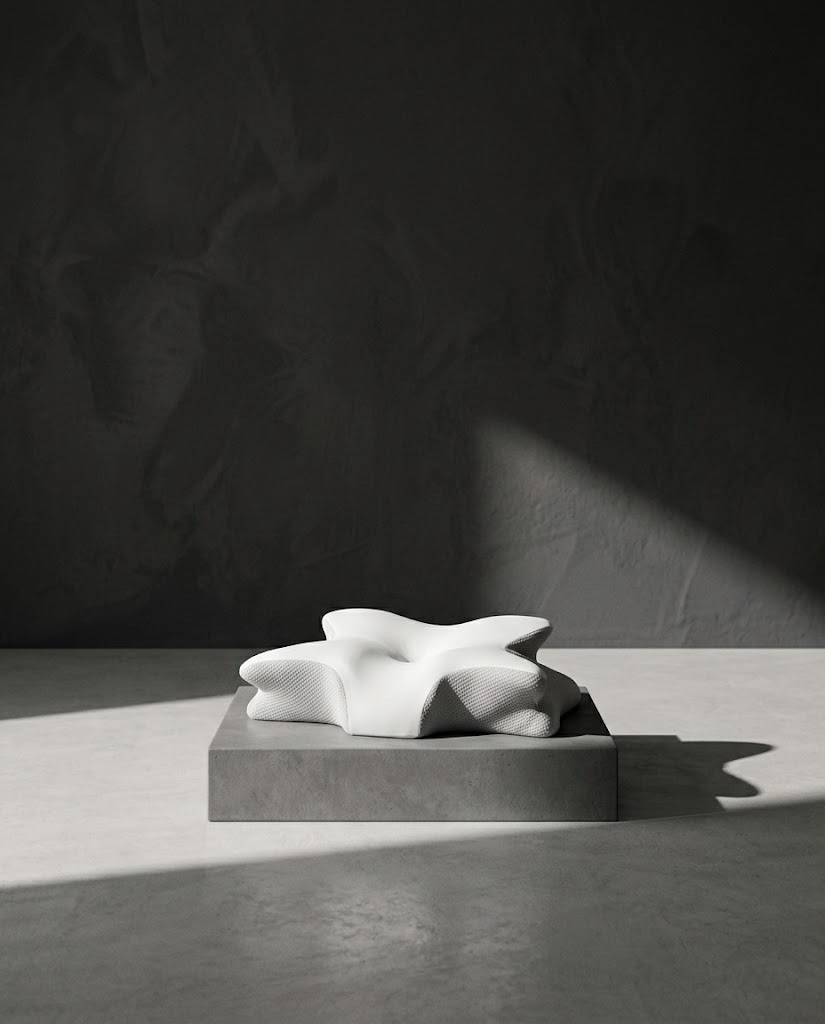 L’oreiller papillon posé sur un socle de béton devant un mur noir texturé, traversé par un rai de lumière en diagonale.